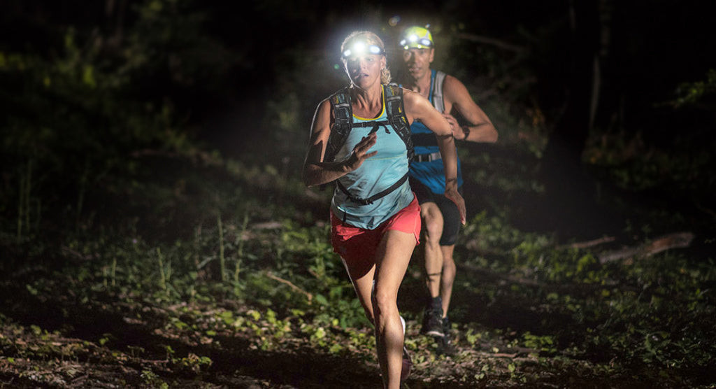 A female and male running on a trail at night in the dark. Both wearing FLEXIT Headlamps and backpacks.