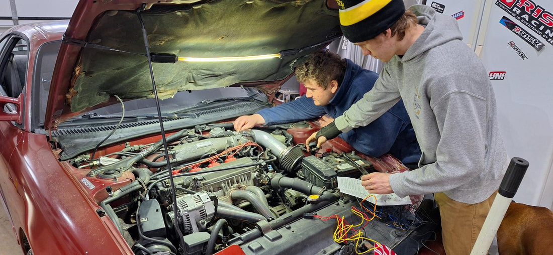 Two young men work together under the hood of a red car, using a FLEXIT Underhood Light mounted to the hood liner for bright, hands-free illumination while working on the engine.
