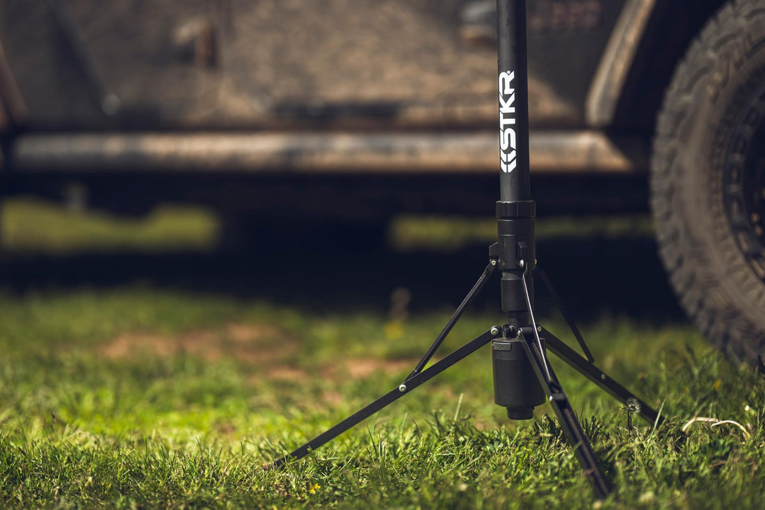 Close up on the base of a FLi OVER-LANDER Telescoping Light sitting in grass with the bottom of an off-road vehicle in the background.