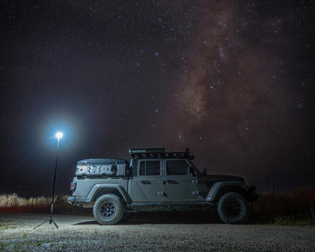A rugged off-road Jeep Gladiator is parked under a star-filled night sky with the Milky Way clearly visible. Next to the vehicle, the FLi OVER-LANDER telescoping area light stands fully extended, brightly illuminating the foreground and part of the Jeep.