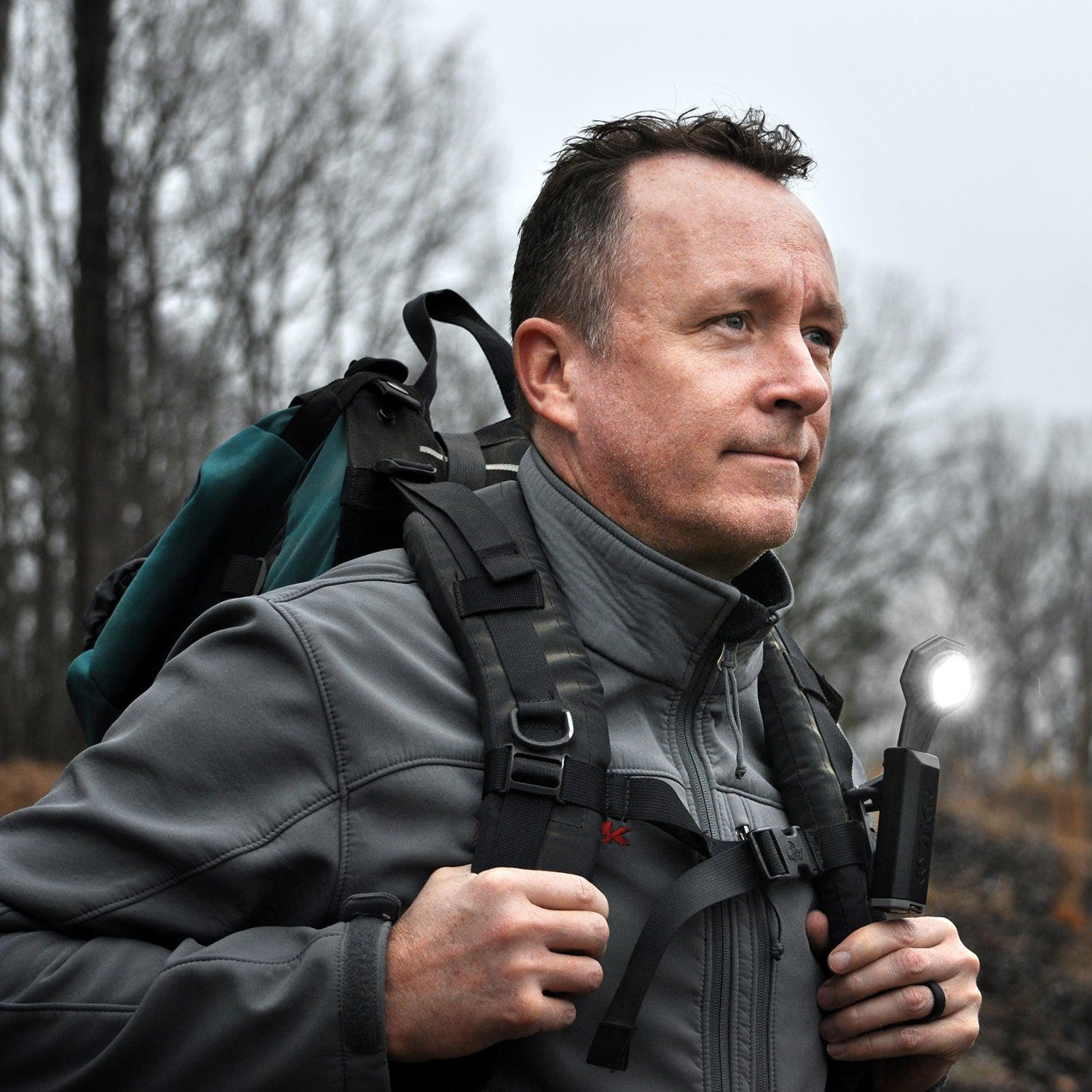 Male hiker in a woods setting wearing a backpack. STKR FLEXIT Pocket Light hooked to one of the backpack straps.