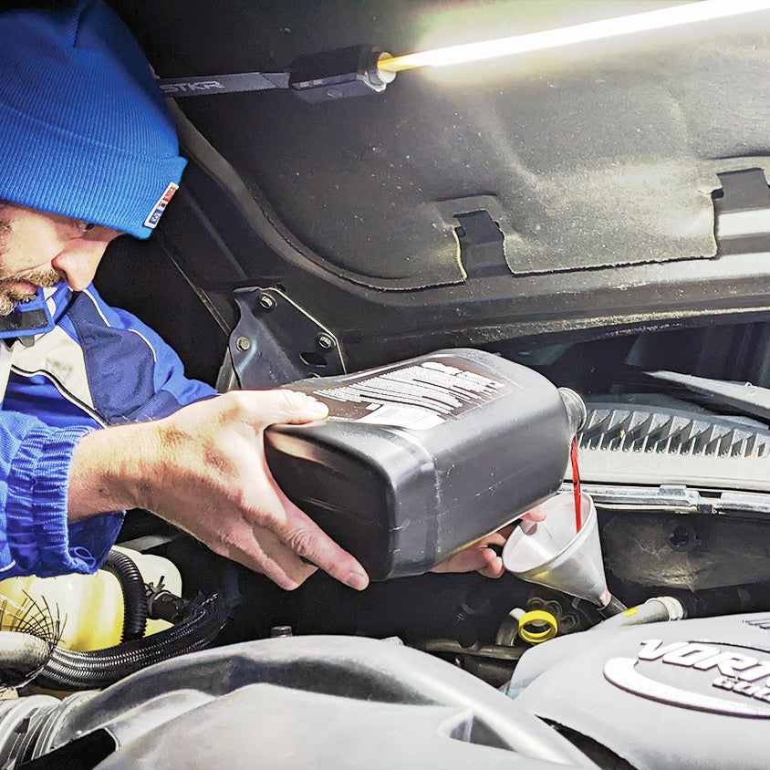 FLEXIT Under Hood Light use image showing a male pouring transmission fluid into a vehicle via a metal funnel. Night time scene light up by an Under Hood Light attached to the underside of the hood.