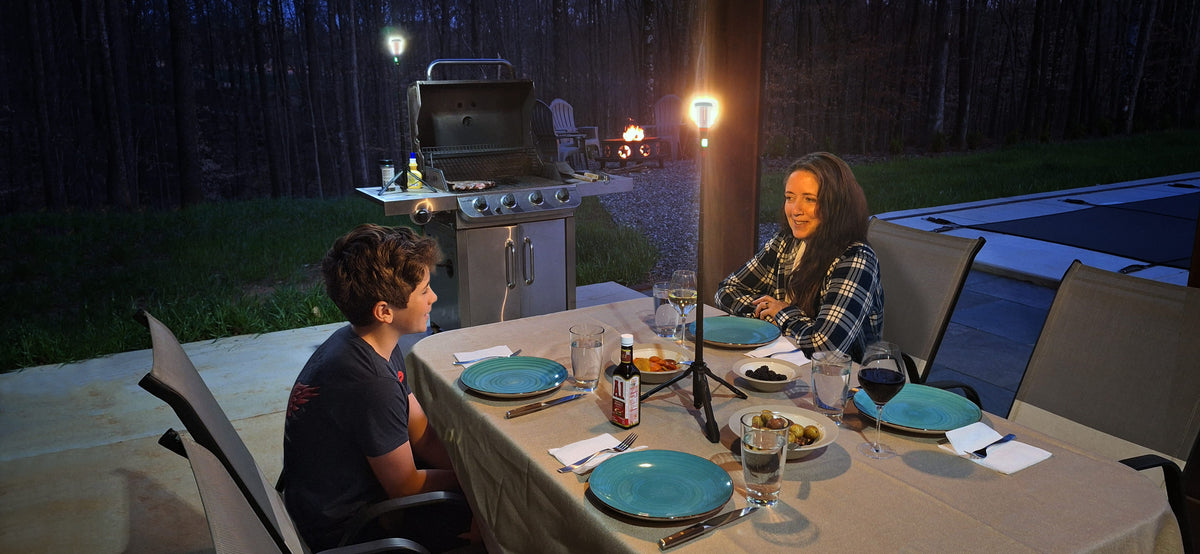 Two people sitting at a table outdoors with plates and glasses, grill in the background. Tabletop telescoping lanterns are sitting on both the table and grill.