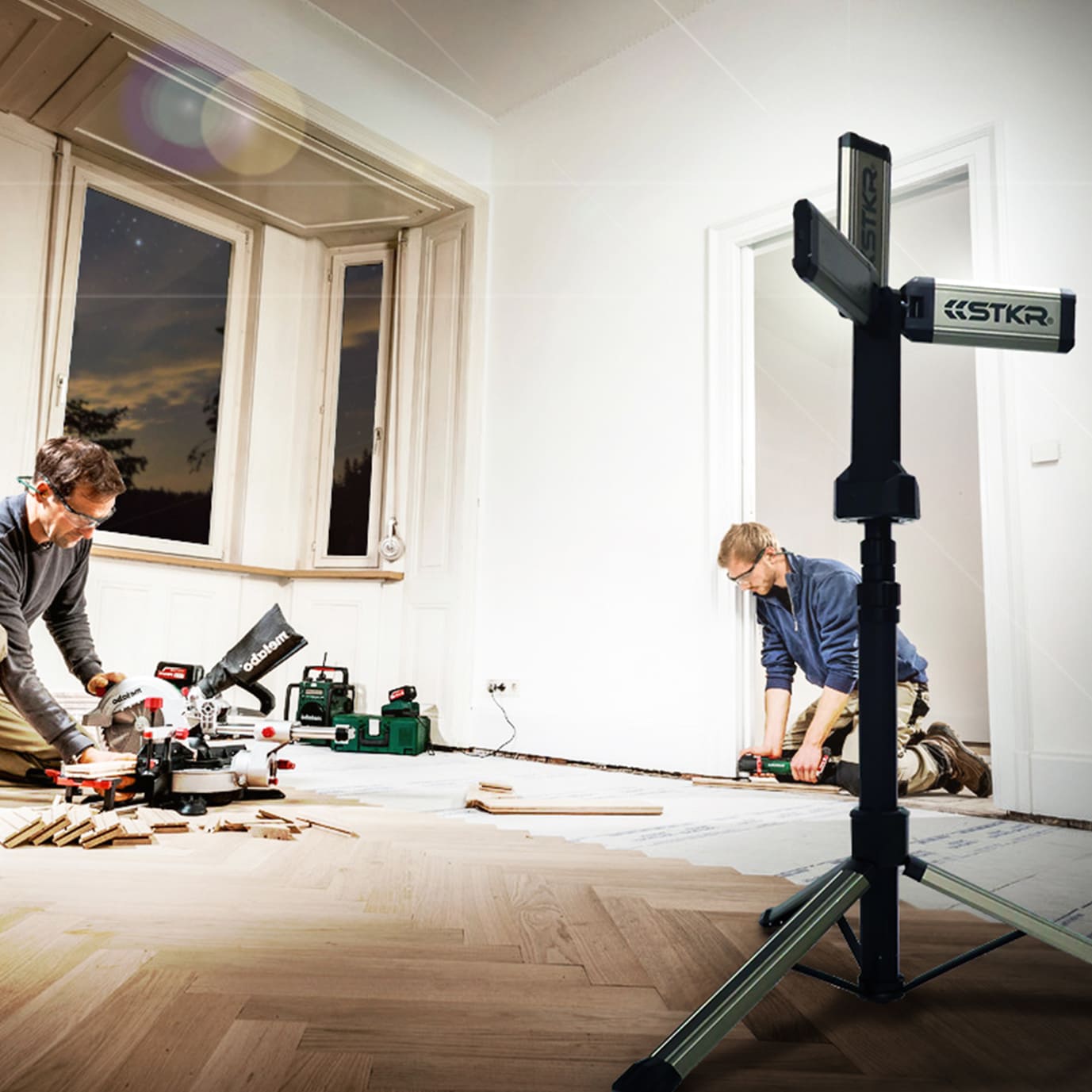 two males installing wood flooring. their work area lit up by an STKR TRi-Mobile on a tripod.